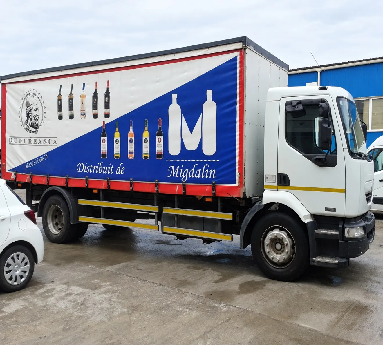 Camion Migdalin cu logo-ul cu sticle și „Distribuit de Migdalin" pe prelată, lângă sticle de vin