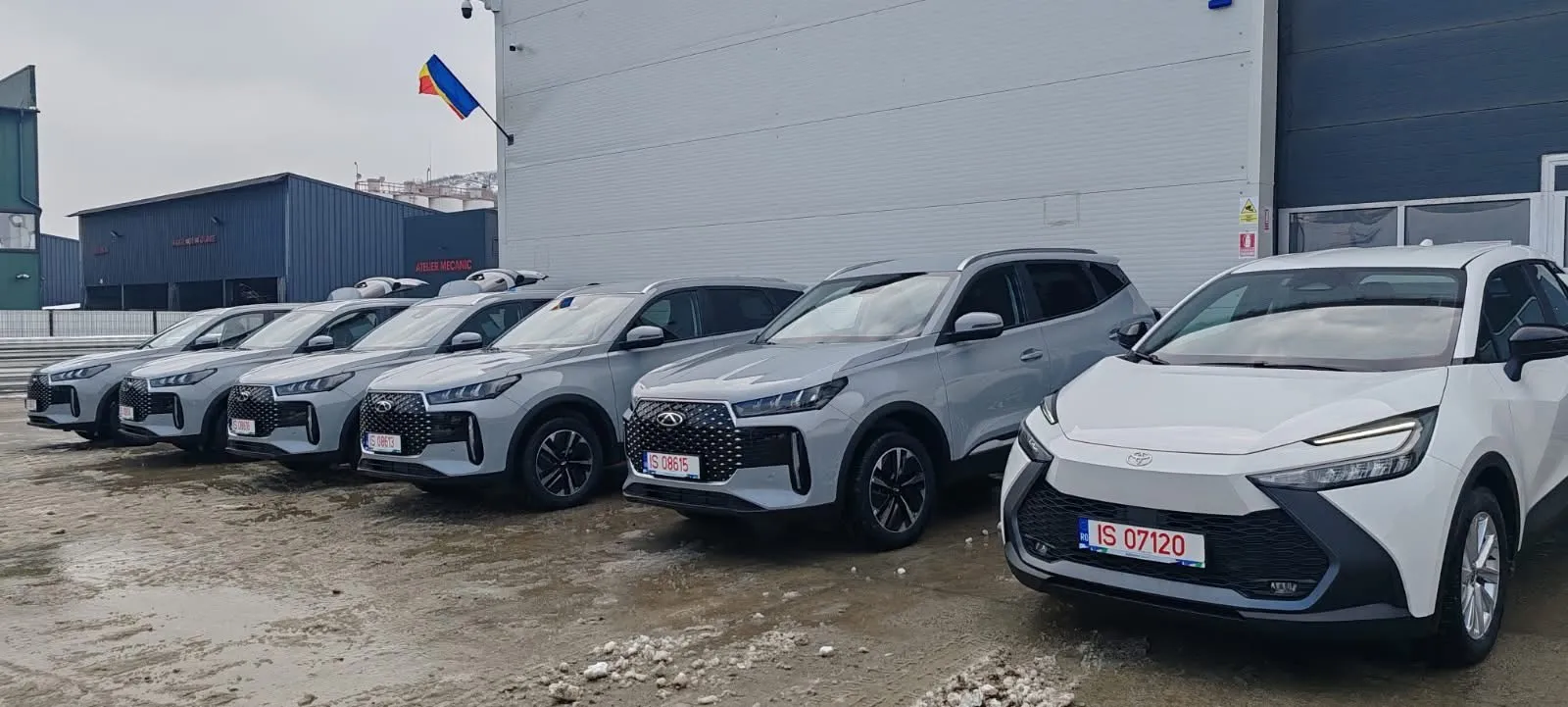 Șase mașini Chery gri și o Toyota albă din flota Migdalin, parcate în fața unei hale