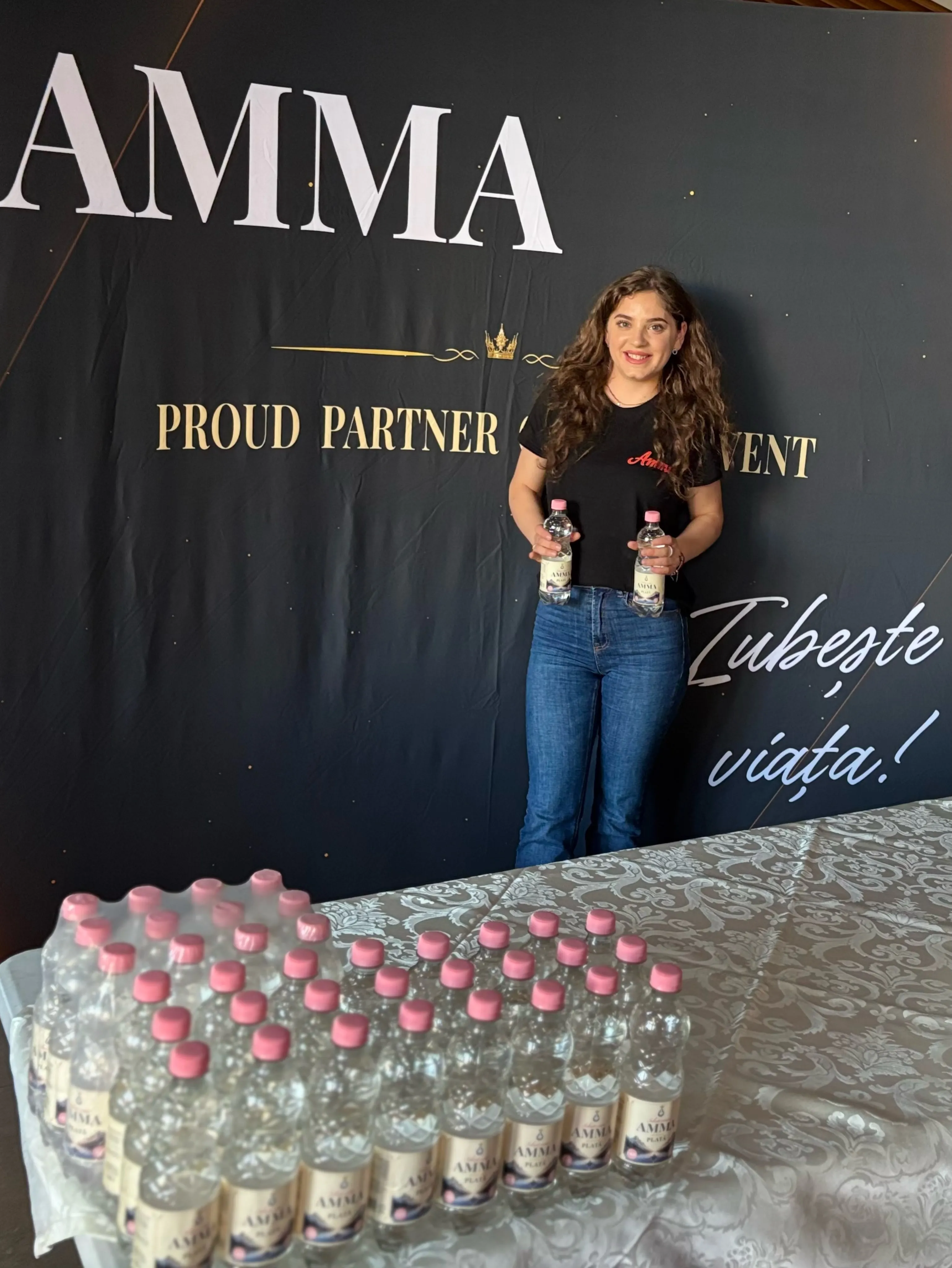 Reprezentantă Amma cu apă Amma, în fața panoului „Proud partner of this event”