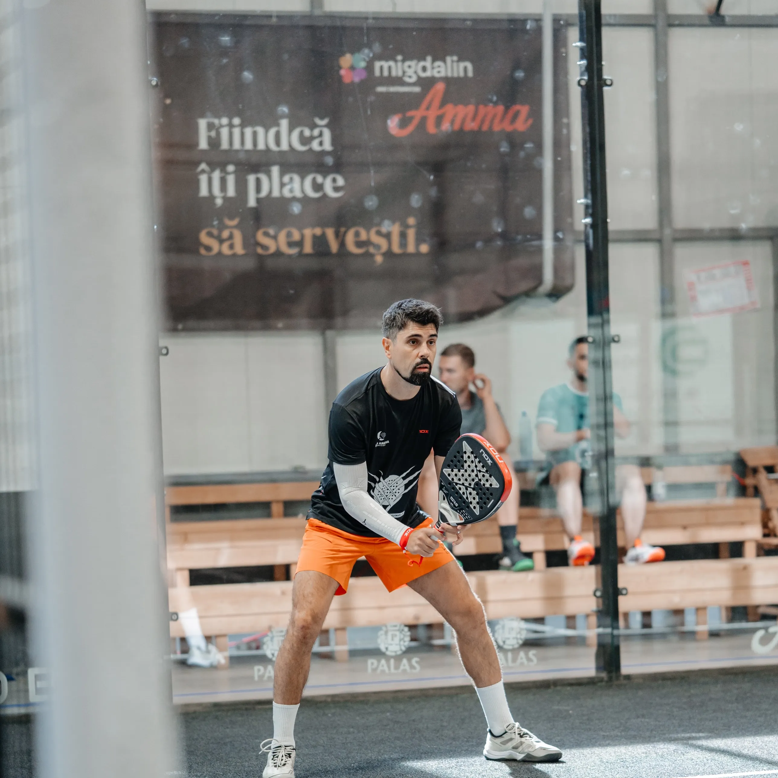 Jucător de padel pe teren, cu bannerul Amma în fundal