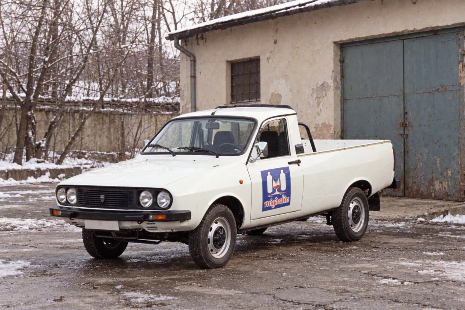 Reconstituire, nu fotografie de epocă. O Dacia Papuc albă cu logo-ul Migdalin cu pahar pe portieră, într-o curte de depozit, iarna